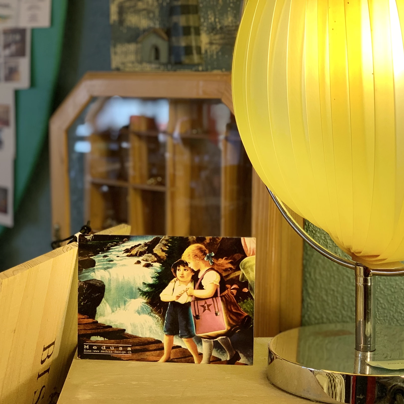 Zu sehen ist der Ausschnitt eines Brockenhauses, wobei im Zentrum eine gelbe Nachttischlampe und einer Postkarte zu sehen sind, im Hintergrund wiederum ein Setzkasten und weitere Gegenstände.