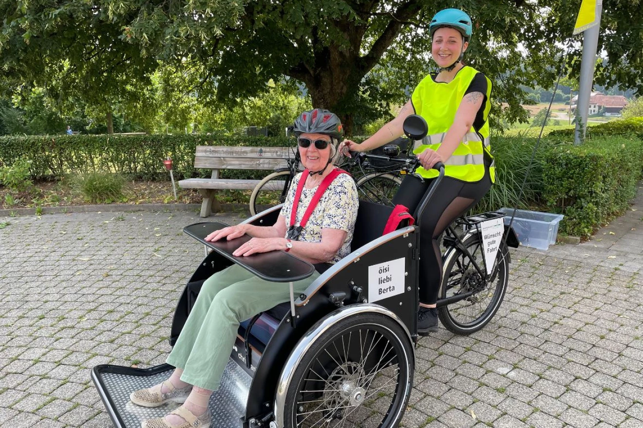 Das Bild zeigt eine Freiwillige auf einer E-Rikscha gemeinsam mit einer Bewohnerin eines Pflegeheims.