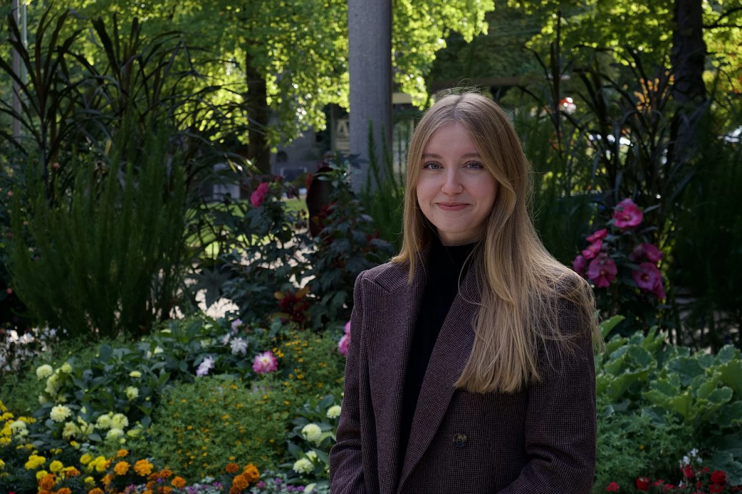 Das Bild zeigt eine freundlich lächelnde junge Freiwillige in einem Park mit vielen bunten Blumen und Bäumen.
