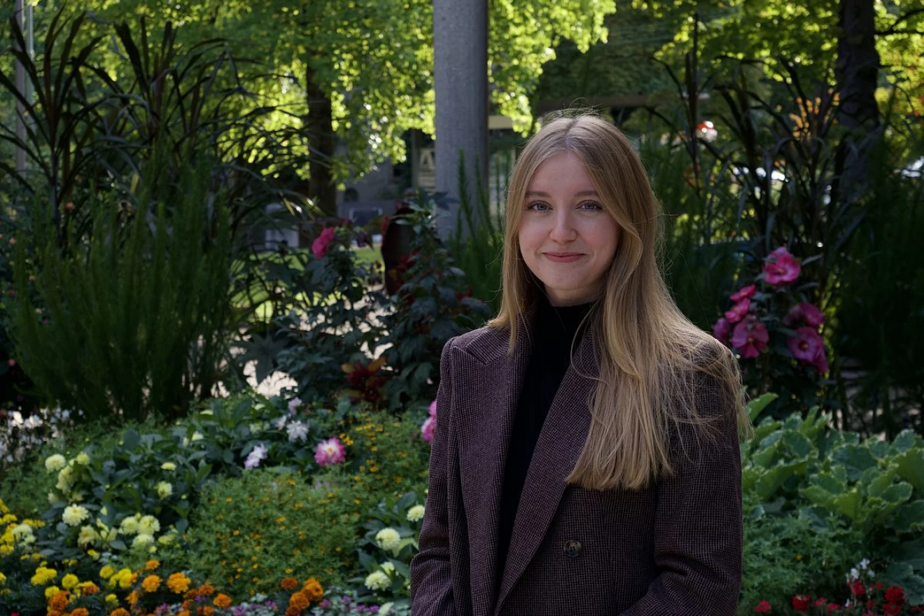 Das Bild zeigt eine freundlich lächelnde junge Freiwillige in einem Park mit vielen bunten Blumen und Bäumen.
