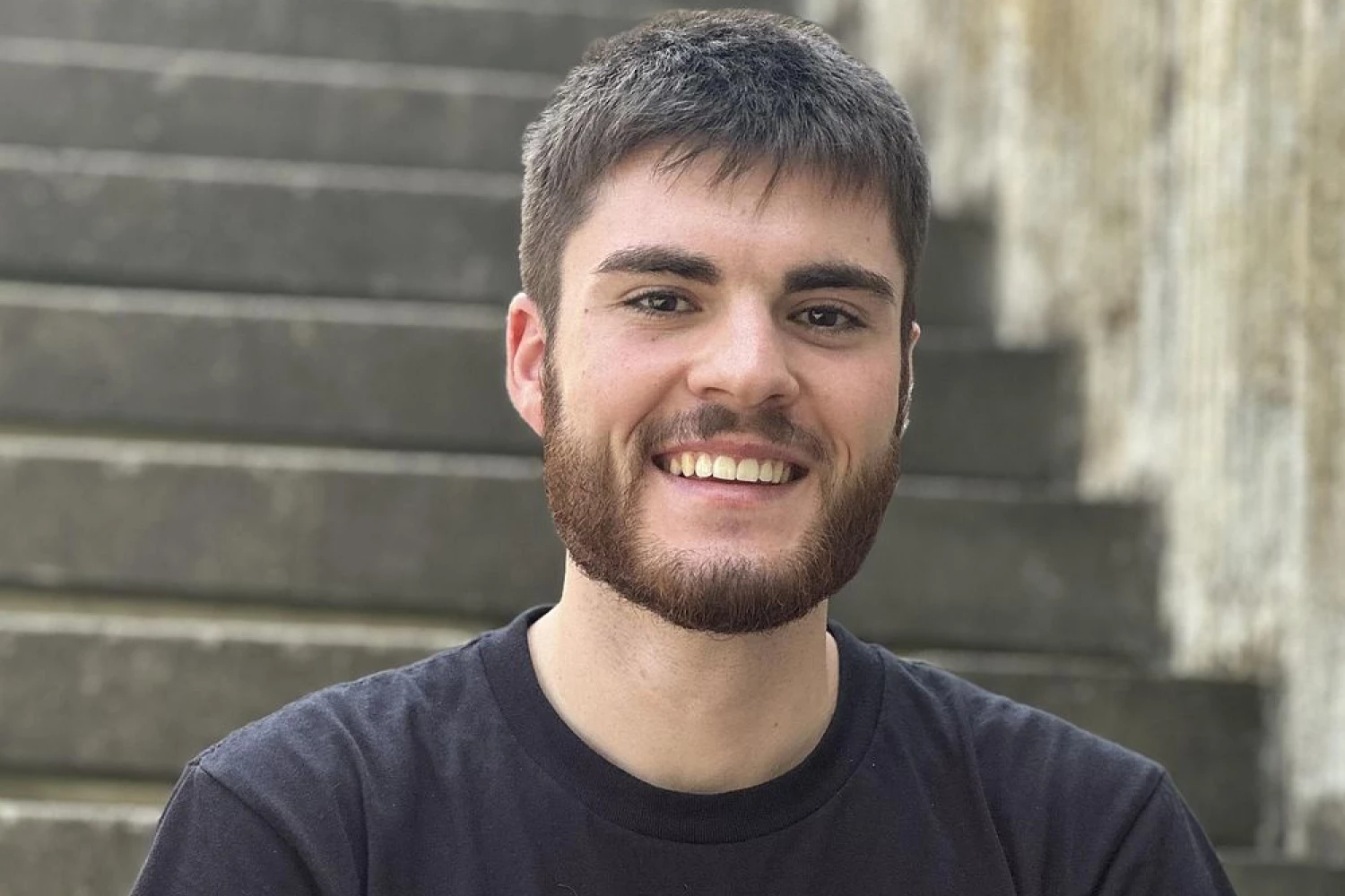 Portrait Foto eines jungen Freiwilligen, der im Freien auf einer Treppe sitzt und freundlich in die Kamera läüchelt.