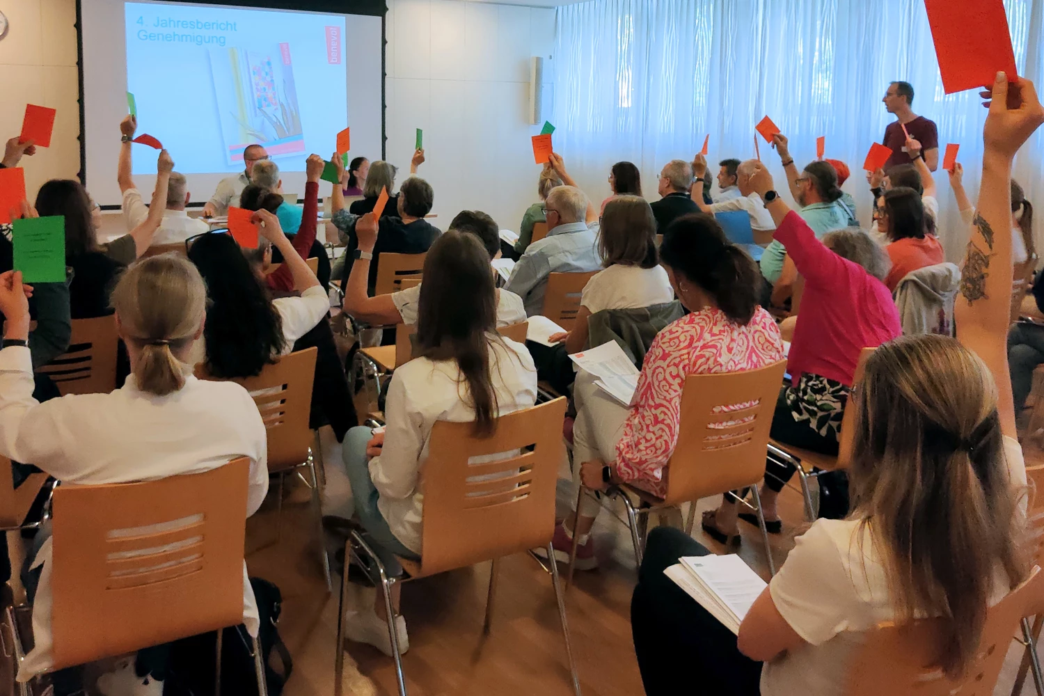 Ca. dreissig Personen sitzen in Konzertbestuhlung mit Blick zu einer Powerpoint-Präsentation und heben orange und grüne Zettel in die Luft.