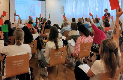 Ca. dreissig Personen sitzen in Konzertbestuhlung mit Blick zu einer Powerpoint-Pr&auml;sentation und heben orange und gr&uuml;ne Zettel in die Luft.